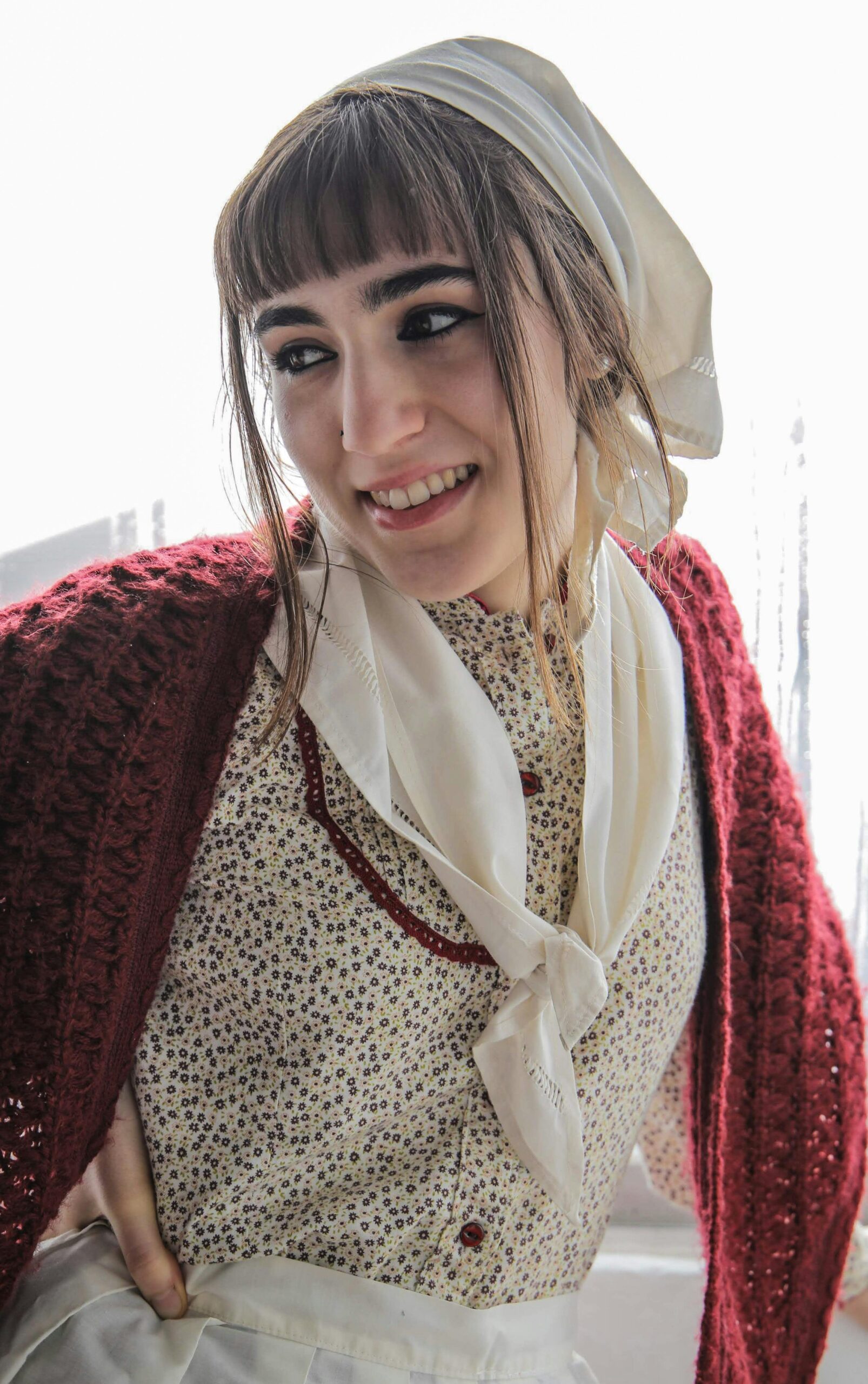 Woman in vintage attire and headscarf smiles warmly indoors. Nostalgic vibe.